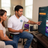A man in a white polo shirt with a TV technician logo sits beside a woman in a living room, pointing at a television screen showing video streaming options. A TV remote, a small speaker, and a purple product box sit on a table nearby.