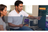 A man in a white polo shirt with a circular logo points toward a television screen showing streaming interface options, while a woman sits beside him in a living room setting.