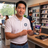 A smiling man in a white polo shirt with a blue circular logo stands in a retail store, holding a smartphone that displays a video or app interface. Shelves with boxed products and other store fixtures are visible in the background.
