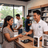 A smiling retail employee in a white polo shirt assists a woman at a counter while she receives a smartphone. An iPhone retail box is visible on the counter, along with store shelves and other staff in the background.