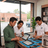 Three people in a retail or service office setting gathered around a table with tablets, phones, and a display box labeled 'Device Warranty Claim' near a large window.